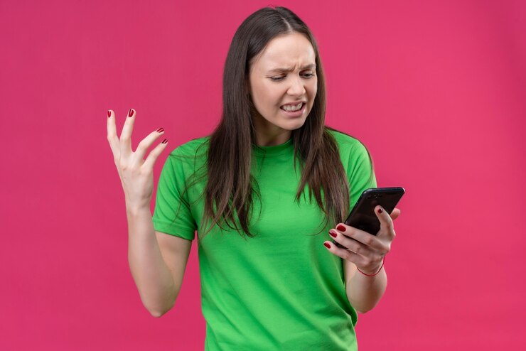 Foto grátis linda garota vestindo uma camiseta verde segurando um smartphone olhando para a tela decepcionada e se sentindo irritada em pé sobre um fundo rosa isolado