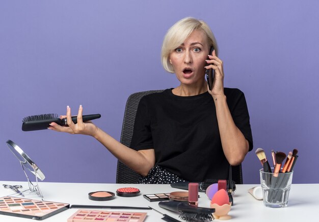 Linda garota zangada se senta à mesa com ferramentas de maquiagem falando no telefone, segurando o pente isolado na parede azul