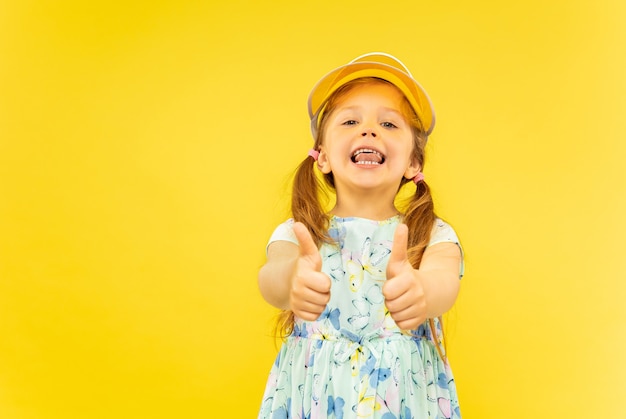 Linda garotinha emocional isolada em fundo amarelo. Retrato de meio comprimento de criança feliz com um vestido e boné laranja mostrando um gesto de OK. Conceito de verão, emoções humanas, infância.