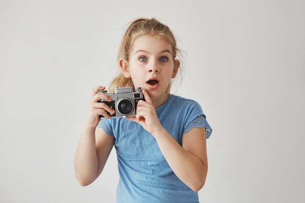 Linda garotinha engraçada com cabelos loiros e olhos azuis, segurando a câmera fotográfica nas mãos, olhando diretamente com expressão assustada.