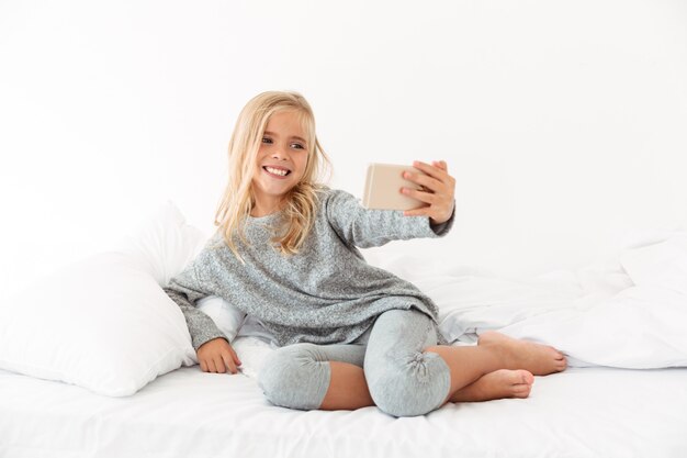 Linda garotinha sorridente tomando selfie no smartphone enquanto estava deitado na cama macia