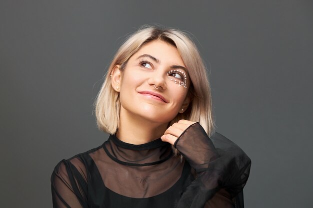 Linda incrível jovem loira com vestido transparente, olhando para cima com uma expressão facial sonhadora, sorrindo misteriosamente, fazendo um desejo.