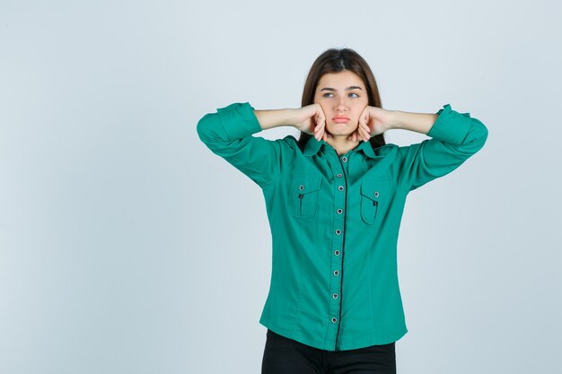 Foto grátis linda jovem amuada com as bochechas apoiadas nas mãos na camisa verde e olhando para baixo, vista frontal.