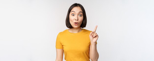 Linda jovem asiática apontando o dedo para cima sorrindo e olhando divertido para a câmera mostrando anúncio de anúncio no fundo branco superior