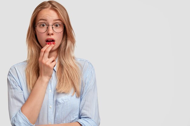 Linda jovem chocada com expressão facial estupefata, mantém a boca aberta, perguntando-se pelas últimas notícias, vestida com uma camisa elegante