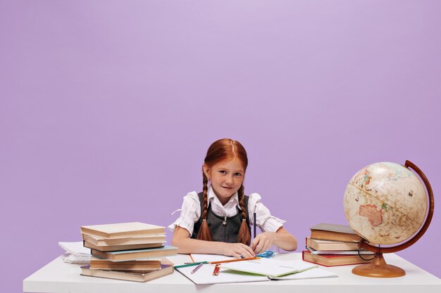 Linda jovem colegial com cabelo vermelho em uniforme escolar, olhando para frente, desenhando e sentada à mesa na parede isolada