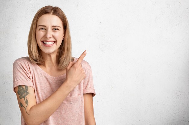 Linda jovem com expressão positiva tem tatuagem, sorri agradavelmente e indica um espaço em branco para o seu texto promocional ou conteúdo publicitário,