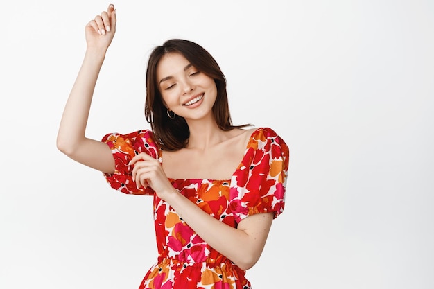 Linda jovem dançando em vestido floral sorrindo e parecendo feliz em pé sobre fundo branco