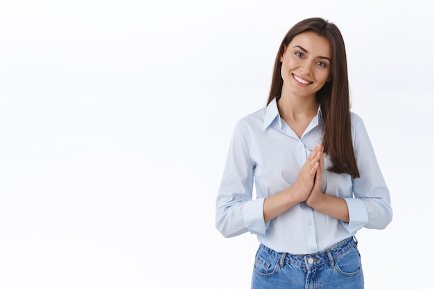 Linda jovem de aparência amigável pronta para ajudar o cliente em qualquer problema, aperte as mãos perto do peito e incline a cabeça, ouvindo com prazer, sorrindo, diga por favor ou agradecendo o favor