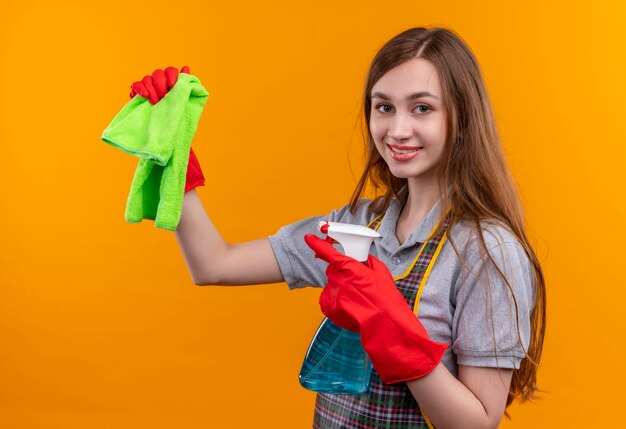 Linda jovem de avental e luvas de borracha segurando spray de limpeza e tapete olhando para a câmera sorrindo, pronta para limpar