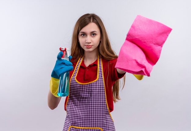 Linda jovem de avental e luvas de borracha segurando spray de limpeza e tapete olhando para a câmera sorrindo, pronta para limpar