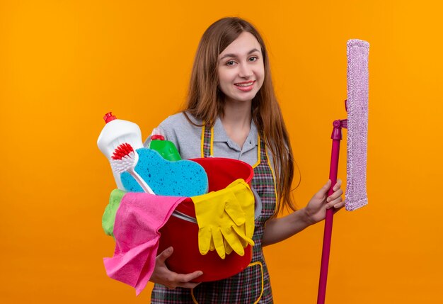 Linda jovem de avental segurando um balde com ferramentas de limpeza e esfregão sorrindo alegremente