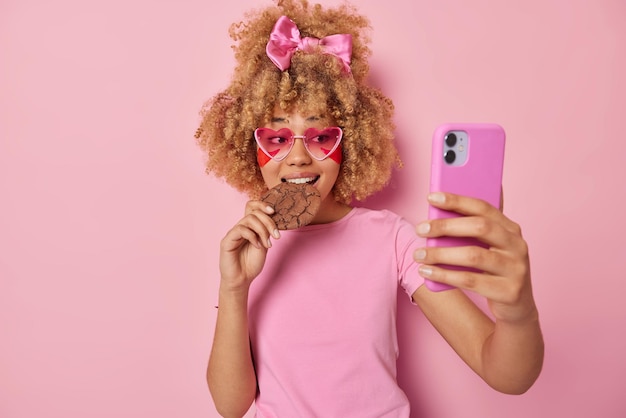 Linda jovem de cabelos encaracolados morde biscoito de chocolate tira selfie via smartphone usa óculos de sol de coração camiseta casual gravata borboleta passa por procedimentos de beleza aplica manchas sob os olhos