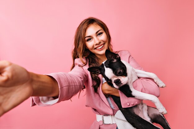 Linda jovem de jaqueta rosa tirando foto de si mesma com o cachorro. Linda garota caucasiana brincando com o bulldog.