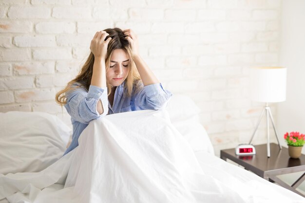 Linda jovem hispânica segurando o cabelo e se sentindo estressada e preocupada enquanto está sentada em uma cama em casa