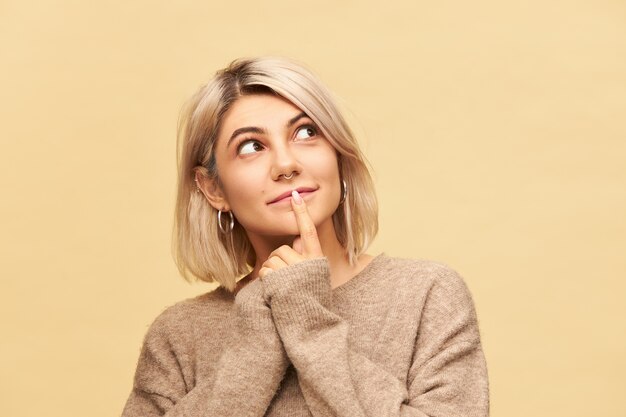 linda jovem linda vestida com um pulôver de cashmere enorme, tendo uma expressão facial curiosa e interessada, olhando para cima, segurando o dedo indicador no lábio, sorrindo. Linguagem corporal