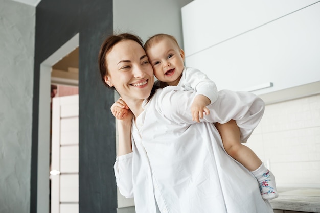 Foto grátis linda jovem mãe brincando com o filho, segurando-o nas costas com as mãos. criança sorrindo brilhantemente e se sentindo feliz.