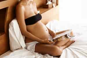 Foto grátis linda jovem mãe expectante caucasiana vestindo blakc cropped top sentado em sua cama com linho branco e lendo o artigo sobre gravidez na revista feminina enquanto descansava dentro de casa em dia ensolarado