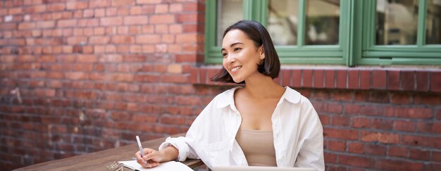 Foto grátis linda jovem modelo sentada com laptop em um café ao ar livre tomando notas escrevendo em seu diário