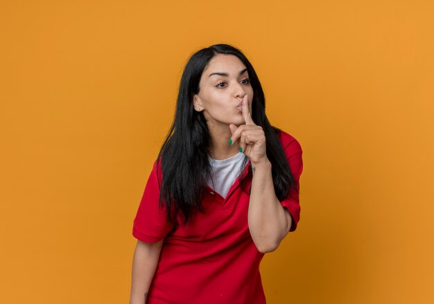 Linda jovem morena caucasiana coloca o dedo na boca gesticulando para sinal de silêncio, vestindo camisa vermelha isolada na parede laranja