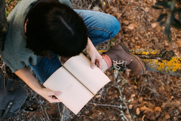 Linda jovem morena sentada nas folhas de outono caídas em um parque, lendo um livro