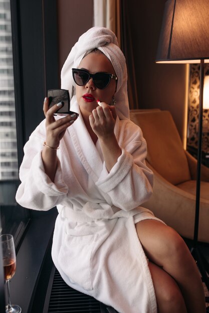Linda jovem mulher branca com cabelo loiro em óculos de sol, vestido roxo, vestido preto, senta-se em seu quarto aconchegante e maquiavel.