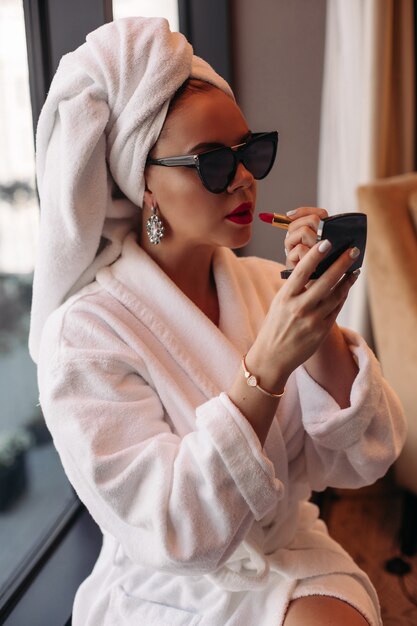 Linda jovem mulher branca com cabelo loiro em óculos de sol, vestido roxo, vestido preto, senta-se em seu quarto aconchegante e maquiavel.