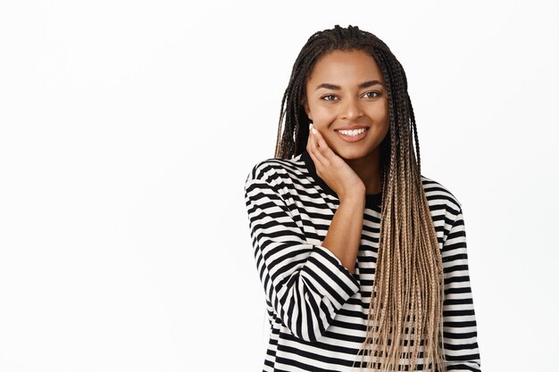 Linda jovem negra com pele facial natural sem maquiagem tocando seu rosto e sorrindo com prazer em pé sobre fundo branco