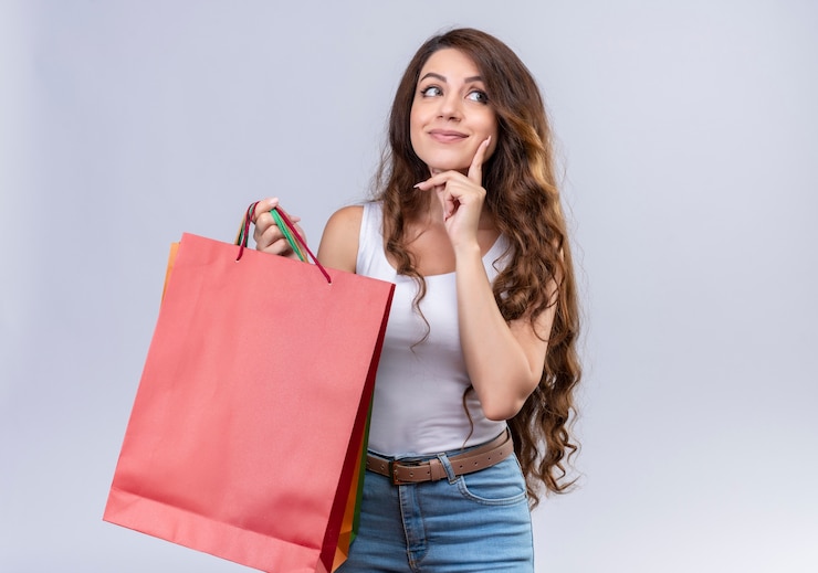 Foto grátis linda jovem pensativa segurando sacolas de compras, olhando para o lado direito com o dedo na bochecha na parede branca isolada