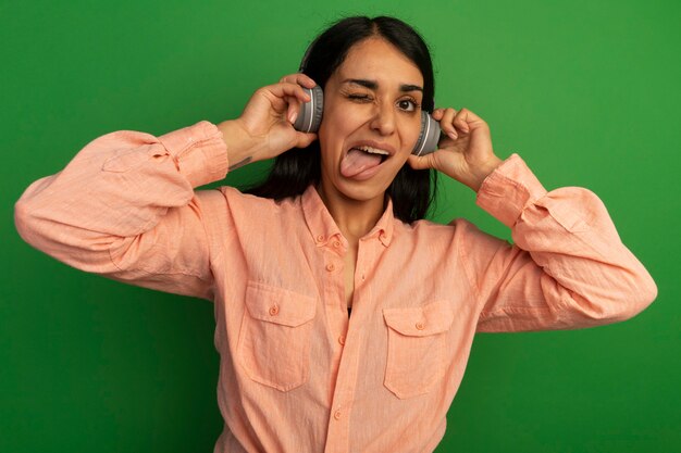 Linda jovem piscando com uma camiseta rosa com fones de ouvido mostrando a língua isolada na parede verde