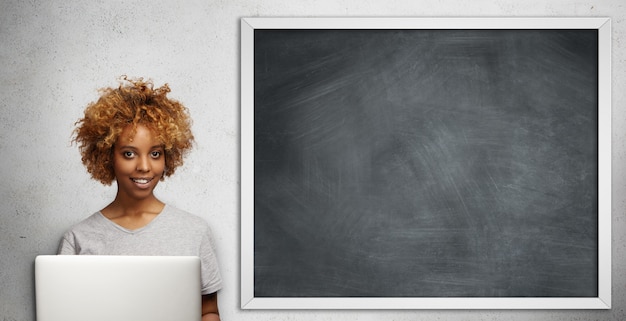 Linda jovem professora de pele escura com um corte de cabelo estiloso, verificando os papéis dos alunos, usando o laptop na sala de aula