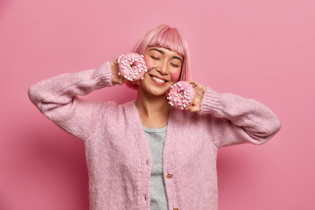 Linda jovem satisfeita sorri com os olhos fechados, segura deliciosos donuts com glacê, imagina o sabor agradável da sobremesa doce, tem o cabelo tingido de rosa, usa um agasalho quente, se diverte, posa em ambientes internos.