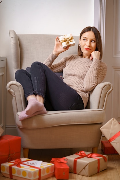 Linda jovem sentada em uma poltrona confortável segurando uma caixa de presente rodeada de presentes em casa.