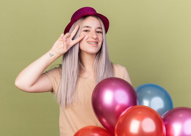 Foto grátis linda jovem sorridente usando aparelho dentário com chapéu de festa em pé atrás de balões, mostrando um gesto de paz