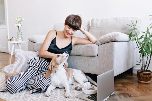 Linda jovem usando calças listradas e relógio de pulso posando no chão enquanto brinca com o cachorro beagle