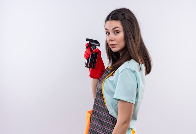 Linda jovem vestindo avental e luvas de borracha segurando um spray de limpeza, de pé de lado com cara séria, olhando para a câmera sobre fundo branco