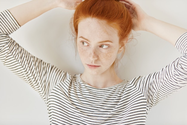 Linda jovem vestindo uma blusa listrada, amarrando o cabelo ruivo em um rabo de cavalo, se preparando antes de ir para a faculdade. Linda ruiva adolescente ganha sardas ajustando o penteado em casa