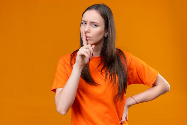 Linda jovem vestindo uma camiseta laranja fazendo gesto de silêncio com o dedo nos lábios, olhando para a câmera com o rosto carrancudo em pé sobre um fundo laranja isolado