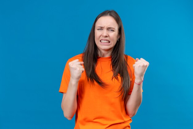 Linda jovem vestindo uma camiseta laranja frustrada, cerrando os punhos e se sentindo irritada em pé sobre um fundo azul isolado