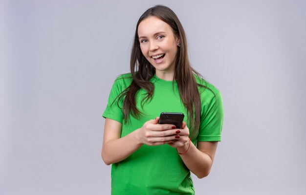 Linda jovem vestindo uma camiseta verde segurando um smartphone, olhando para a câmera, sorrindo alegremente em pé sobre um fundo branco isolado