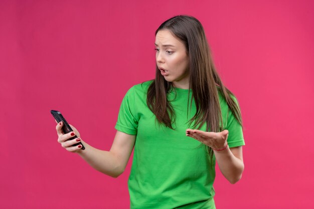 Linda jovem vestindo uma camiseta verde segurando um smartphone, olhando para a tela, confusa e muito ansiosa em pé sobre um fundo rosa isolado