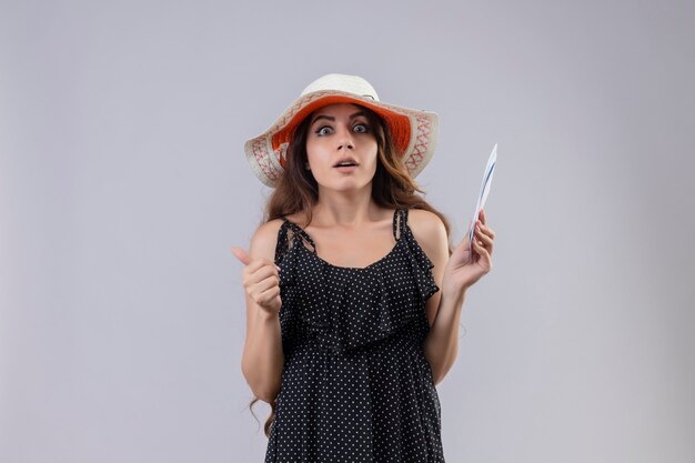 Linda jovem viajante com um vestido de bolinhas e um chapéu de verão segurando as passagens aéreas, parecendo chocada e assustada em pé sobre um fundo branco