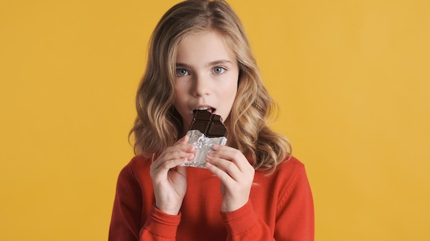 Linda loira adolescente comendo barra de chocolate isolada em fundo amarelo. menina de dente doce