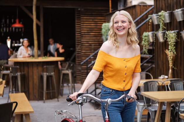 Linda loira alegre de pé com bicicleta clássica enquanto descansava alegremente no pátio do café