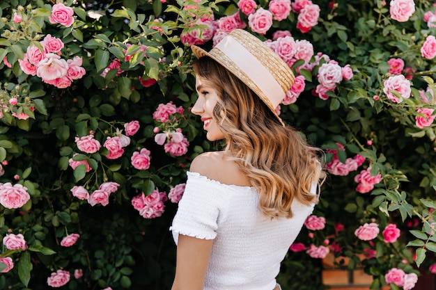 Foto grátis linda loira com chapéu de verão, olhando para as flores com um sorriso. mulher encaracolada satisfeita relaxando durante a sessão de fotos com rosas.