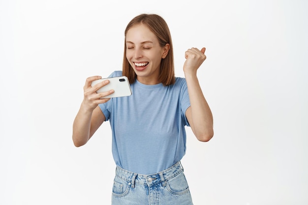 Foto grátis linda loira comemorando a vitória, atingir o objetivo no aplicativo, assistindo a transmissão ao vivo no smartphone, tela invertida horizontal, regozijando-se com a bomba de punho, sorrindo satisfeito, fundo branco.