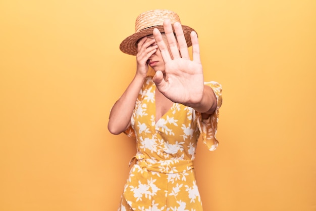 Foto grátis linda loira de férias usando chapéu de verão e vestido sobre fundo amarelo, cobrindo os olhos com as mãos e fazendo gesto de parada com expressão triste e de medo conceito embaraçado e negativo