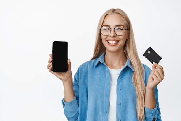 Linda loira de óculos sorrindo mostrando a tela do celular e cartão de crédito demonstrando o aplicativo de finanças sobre fundo branco