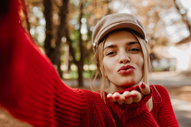 Linda loira faz um beijo em sua selfie no parque. Linda mulher usa um elegante suéter vermelho e um chapéu leve.