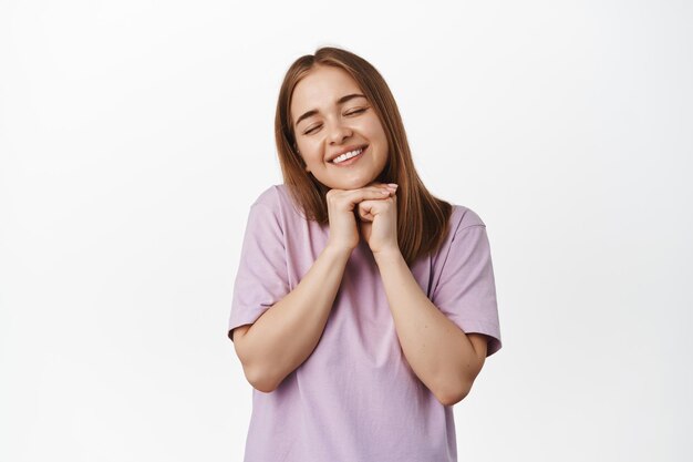 Linda loira fecha os olhos, sorrindo e sonhando, rezando ou fazendo desejos, antecipando algo lindo, sonhando acordado, de pé contra um fundo branco em t-shirt